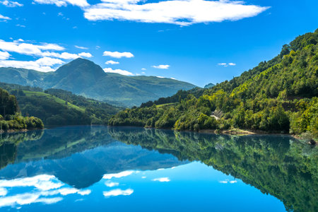 The serene reservoir in Tolosa, Gipuzkoa, reflects the surrounding mountains and blue sky with white...