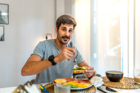 Happy man enjoying avocado toast alongside ham and eggs while relaxing in his modern apartment,...
