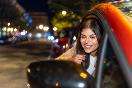 Young woman leaning out of a car window at night, smiling as city lights and bokeh blur by, enjoying...