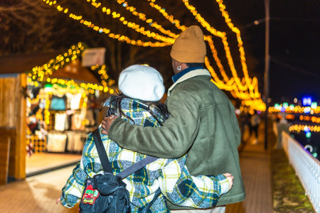Multiethnic couple strolling through a beautifully decorated outdoor Christmas market, embracing the...