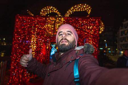 Tourist taking a selfie and making thumbs up sign with christmas lights and decorations in bayonne,...