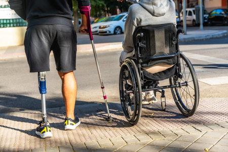 Two people with disabilities moving on a city street, one using a prosthetic leg and a crutch, and...