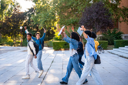 Diverse group of young university students walking on campus after classes, happily raising arms in...