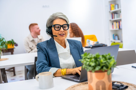 Senior woman actively working in a modern office, wearing a headset and glasses while typing on a laptop, engaging in customer service or remote communication in a collaborative workspaceの写真素材