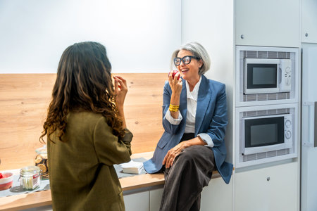 Two business women colleagues having a healthy snack, eating apples and laughing together while...