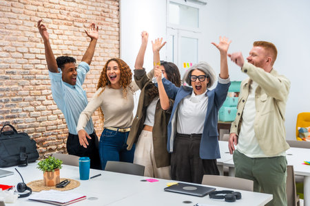 Multiracial business team in a modern office cheering with arms raised, celebrating a shared success...
