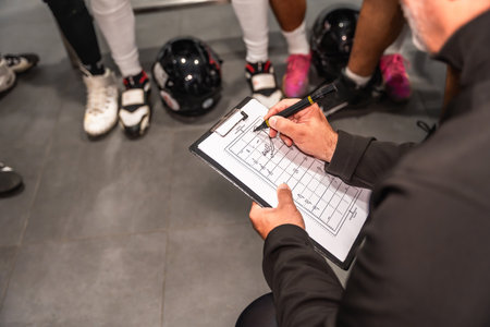 American football coach holding a clipboard, drawing strategies on a field diagram, instructing players in a locker room during a huddle, discussing tactics and game planの写真素材
