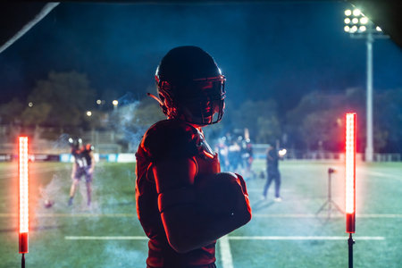 American football player in full gear holding a ball, standing confidently on a floodlit stadium field at night, intensely focusing on the upcoming game or practiceの写真素材