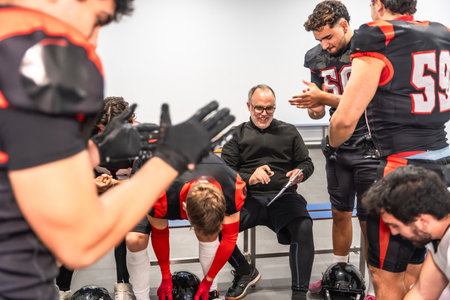 American football coach smiling, discussing strategy, and preparing for a game with diverse male players dressed in uniforms and protective gear inside a locker roomの写真素材