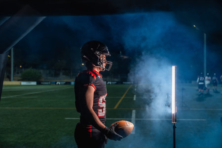 American football player is holding a ball, wearing a helmet and uniform, and standing on a playing...