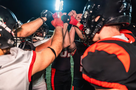 American football players huddling on the illuminated field, placing their hands together in the center, demonstrating teamwork, unity, and determination during a night gameの写真素材