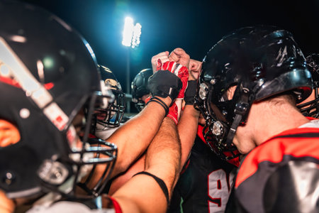 American football players putting hands together in a huddle, symbolizing teamwork, unity, strategy, and motivation on a field during a night game under stadium lightsの写真素材