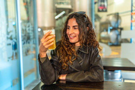 Woman in a leather jacket smiling and savoring a glass of craft beer at a modern brewery bar, enjoying a relaxed tasting experience with friends in a casual weekend hangoutの写真素材