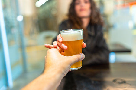 Hand holding a refreshing glass of craft beer, presenting it to a woman in a bar, representing tasting, brewery visits, social gatherings, and enjoying drinksの写真素材