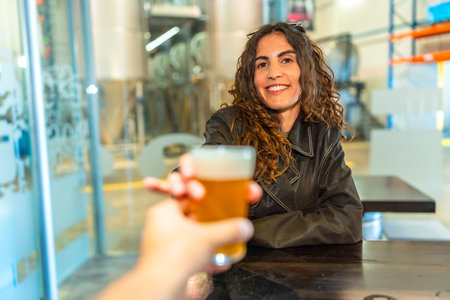 Woman smiling and raising a glass of frothy craft beer in a modern brewery, toasting an unseen companion across a wooden table during a relaxed, social weekend outingの写真素材