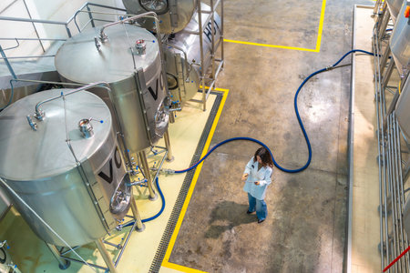 Woman scientist in a white lab coat monitoring stainless steel fermentation tanks in a modern...