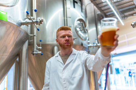 Brewery worker performing quality control by examining a glass of amber craft beer, ensuring production standards and evaluating the beverage for clarity, color, and foam in a factory settingの写真素材