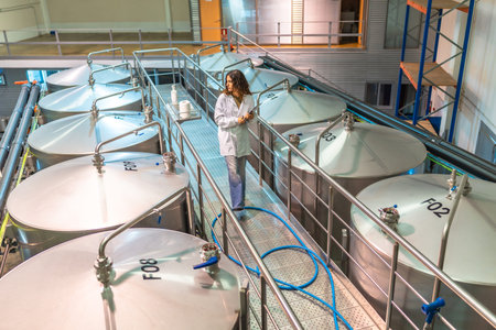 Woman scientist in a lab coat walking on a platform among large industrial fermentation tanks,...