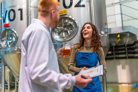 Brewery worker and quality control specialist smiling, holding clipboard and a glass of beer,...