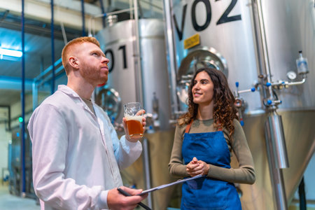 Two brewery professionals inspect and taste craft beer beside large fermentation tanks in a modern microbrewery, checking quality, recording results and collaborating on productionの写真素材