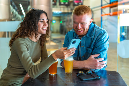 Young couple sitting at a table in a brewery or beer factory, having a conversation and sharing content on a smartphone while enjoying refreshing glasses of craft beerの写真素材