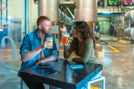 Couple sitting at a table in a modern craft beer factory, drinking and communicating while enjoying a brewery tour experience with large fermentation tanks in the backgroundの写真素材