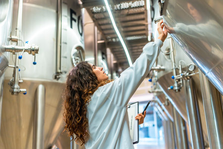 Young woman in a lab coat checking control panel on a stainless steel fermentation tank, performing quality control and inspection during the beer production process at a modern facilityの写真素材