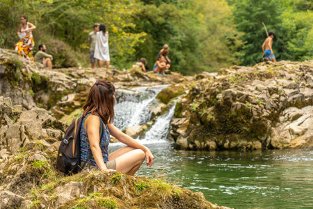 Young female hiker resting on rocks near a small waterfall on the Dobra river, with other hikers enjoying the natural pool in the background, on a sunny day in Asturias, Spainの写真素材