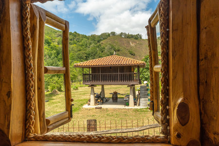 Open wooden window with rope hinges framing a scenic view of a traditional elevated horreo granary in the serene countryside of Asturias, Spainの写真素材