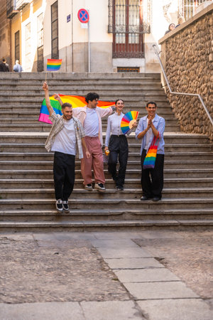 Young friends are celebrating lgbtq plus pride by walking down urban stairs, holding a large rainbow flag, a small flag, and a fan, expressing joy and unityの写真素材