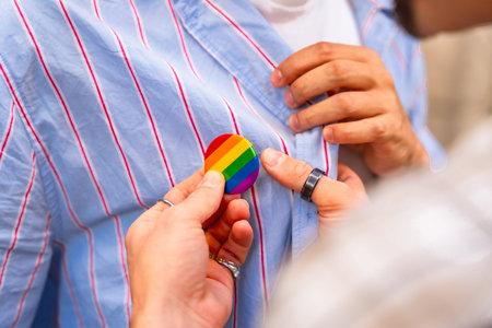 Young couples hands pinning a rainbow pride flag button to a striped shirt, close up symbolizing...