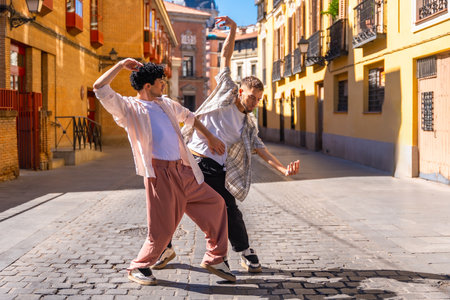 Two young men performing energetic contemporary street dance on cobblestones in madrid, expressing freedom, connection and joy under bright summer sunlight in an urban cityscapeの写真素材