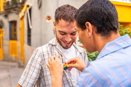 Young gay man gently attaching a rainbow pride flag pin to the plaid shirt of his smiling partner, celebrating love and lgbtq plus rights on a city streetの写真素材