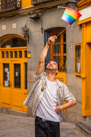 Young man standing on a street, smiling and raising a small rainbow flag, expressing happiness and support for the lgbtq community during a celebration eventの写真素材