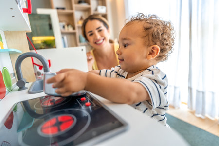 Happy toddler playing with toy kitchen while his mother watches him, promoting early childhood development and imaginative playの写真素材