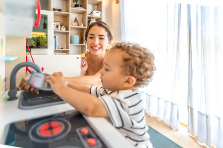 Smiling mother looking at her little son having fun playing with a toy kitchen in a modern, bright living roomの写真素材