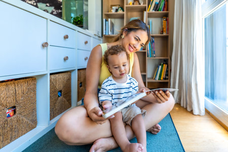Happy mother and baby son reading a book together, sitting on the floor in the living room at homeの写真素材