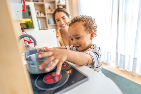 Happy toddler boy having fun playing with a toy kitchen while his mother watches over him in a bright and airy homeの写真素材
