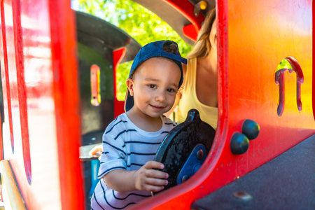 Little boy having fun driving a toy train at the playground, with his mother supervising in the background, enjoying a sunny day outdoorsの写真素材