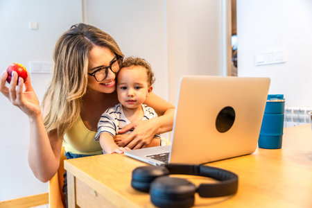 Mother holding an apple while hugging her son, both enjoying a moment together, watching something entertaining on the laptop at homeの写真素材