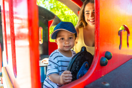 Happy toddler having fun driving a toy train at the playground while his mother watches him with a smileの写真素材