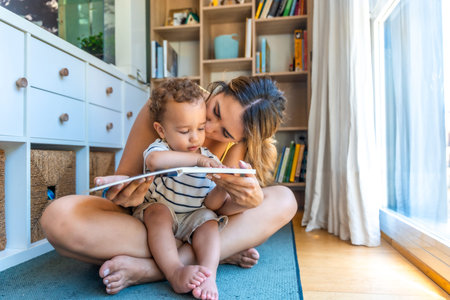 Mom kissing her little son while reading a book together, sitting on the floor in the living roomの写真素材