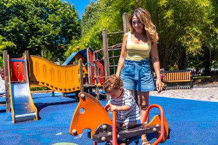 Young mother assisting her son while playing on a rocking horse in a colorful playground on a sunny summer dayの写真素材