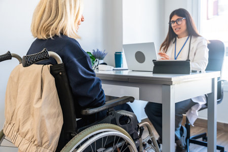 Patient in a wheelchair receiving medical advice from a female doctor during a healthcare consultation in a bright clinic setting, focusing on disability care and supportの写真素材