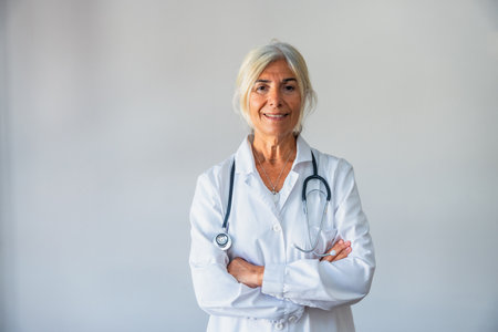 Senior female doctor with gray hair and a lab coat smiling confidently, standing against a neutral background with a stethoscope around her neck, representing healthcare and medical professionalismの写真素材