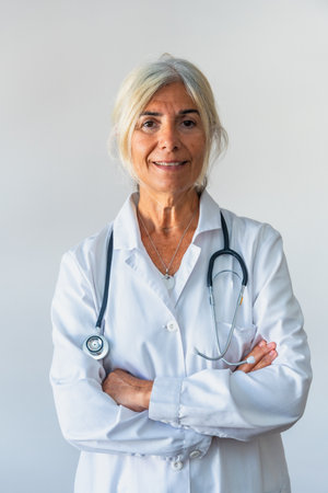 Senior woman doctor with gray hair and lab coat standing confidently with arms crossed, stethoscope around her neck and smiling at camera, conveying experience, trust and professional medical careの写真素材
