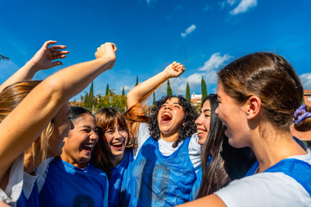 Young women soccer players cheering with arms raised in celebration, smiling and embracing after victory under a blue sky, showing teamwork, joy, friendship and empowermentの写真素材