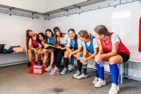 Female athletes wearing sports attire sitting on a bench in a locker room, discussing strategy and reviewing a folder before or after a sports event, showing teamwork and communicationの写真素材