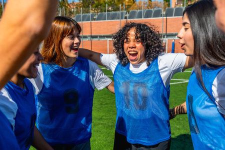 Young diverse women football players joining arms in a huddle, enthusiastically celebrating a...