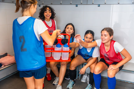 Teenagers from a diverse girls soccer team taking a water break and hydrating, showing friendship, sports, and teamwork in the locker room after practice or a gameの写真素材